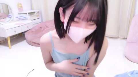 Media: A young Asian woman with straight black hair and pale skin, wearing a light blue camisole, a white face mask, and a pink choker, kneels on a white floor in a cozy room with a pink beanbag, white dresser, and mirror.