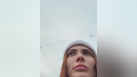 Media: Video of a young woman with fair skin, red hair, and light eyes, wearing a grey beanie, looking upward, with a minimalist white room background.