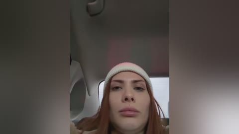 Media: Video of a young woman with fair skin, straight red hair, and a light pink and white beanie, wearing a neutral expression. She sits in a dimly lit car interior, with blurry, out-of-focus background.