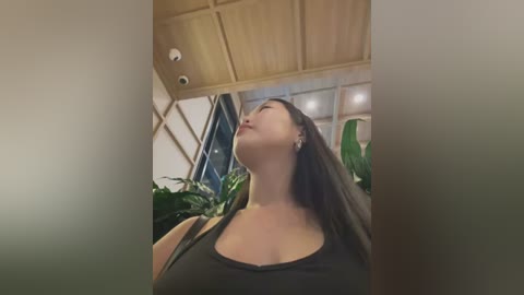 Media: Video of a young Asian woman with long, straight black hair, wearing a black tank top, standing in a greenhouse with lush green plants and wooden beams, looking up with a serene expression.