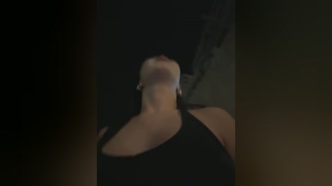 Media: Video of a woman with fair skin and long, dark hair, wearing a low-cut black top, captured from a low angle, emphasizing her cleavage and neck, against a dark, blurry background.