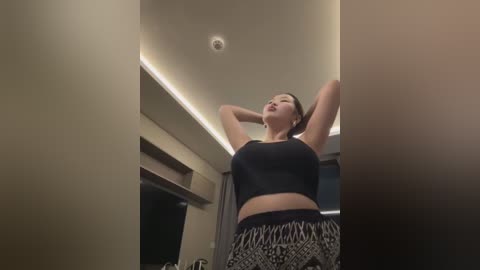 Media: A video of an Asian woman with fair skin and dark hair, wearing a black sports bra and patterned pants, stretching in a modern room with beige walls and a recessed ceiling light.