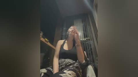 Media: A video of a young woman with fair skin, dark hair, wearing a black tank top and patterned blanket, sitting on a bed with a wooden headboard, her hands covering her face, looking distressed.