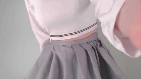 Media: Video of a person wearing a white, long-sleeved sweater with a black ribbon around the waist, paired with a pleated gray skirt. The background is plain and light gray, focusing on the clothing details.