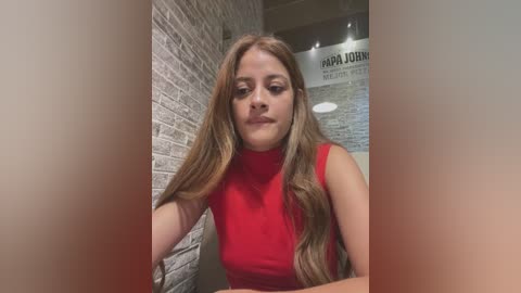 Media: Video of a young woman with long, wavy light brown hair, wearing a sleeveless red top, standing against a brick wall in a dimly lit room with a blurred background.