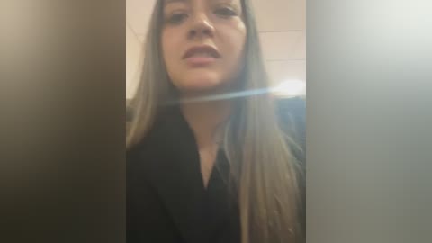 Media: Video of a young woman with long, straight blonde hair, wearing a black jacket, captured from a low angle in a dimly lit room with blurred background.