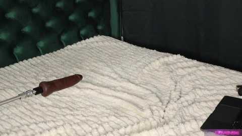 Media: Video of a bed with a tufted, dark green headboard. A cream-colored, textured blanket covers the bed. A large, wooden-handled knife lies on the blanket, positioned diagonally. The image is dimly lit, emphasizing the texture of the blanket and the knife.