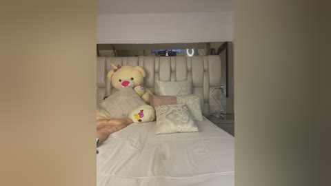 Media: Video of a plush, yellow teddy bear on a white bed with tufted headboard, surrounded by white pillows and a beige blanket.