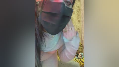 Media: Video of a person wearing a black face mask, a light grey shirt, and a pink mask, standing in a room with a tiger-themed pillow on the floor.