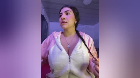 Media: Video of a plus-size Latina woman with long dark hair, wearing a pink and white striped hoodie, standing in a dimly lit room with purple lighting.