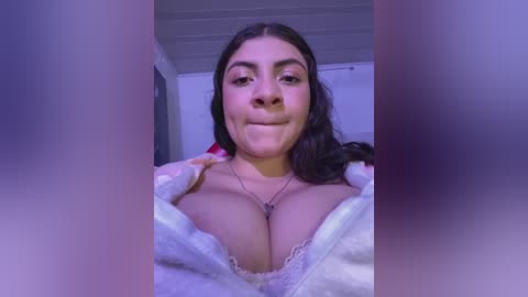 Media: Video of a smiling Latina woman with long dark hair, wearing a white lace bra, revealing ample cleavage, in a dimly lit room with a purple tint.