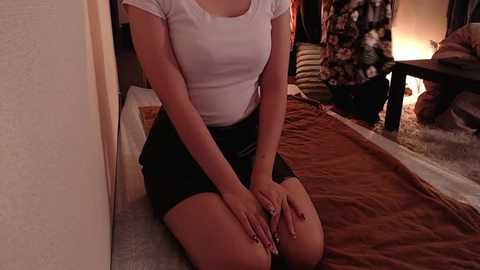 Media: Video of a woman in a white t-shirt and black skirt, kneeling on a brown blanket in a dimly lit room with a fireplace and a floral-patterned couch in the background.