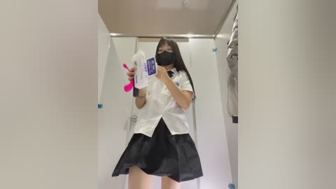 Media: Video of a young East Asian woman with straight black hair, wearing a white blouse, black skirt, and black face mask, holding a pink vibrator in a changing room.