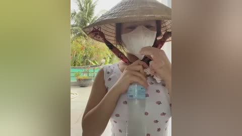 Media: Video of a woman in a white dress with red patterns, wearing a traditional conical hat and a face mask, holding a water bottle outside in a tropical garden.