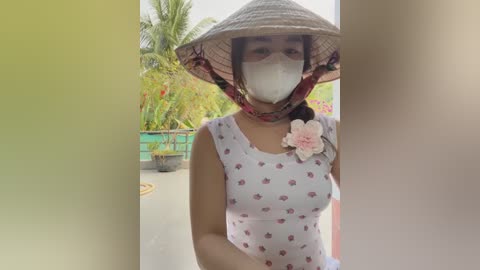 Media: Video of a woman wearing a traditional Vietnamese conical hat, white mask, floral dress, and pink flower brooch, standing in a lush garden with greenery and potted plants in the background.