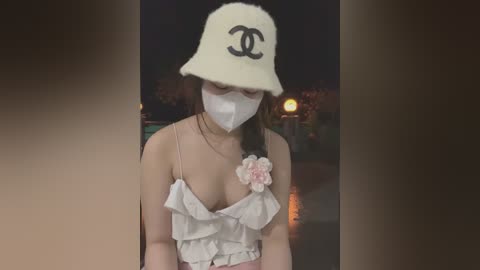 Media: A video of an Asian woman with fair skin, wearing a white Chanel bucket hat, face mask, and a ruffled white crop top with a pink flower, posing outdoors at night.