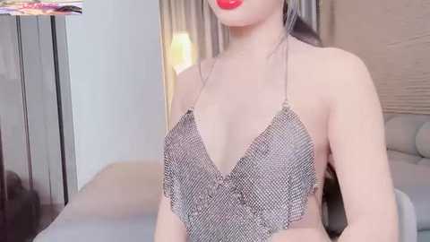 Media: Video of a fair-skinned woman with long black hair, wearing a metallic, beaded halter neck top, standing in a modern, softly lit bedroom with beige walls and a large bed.