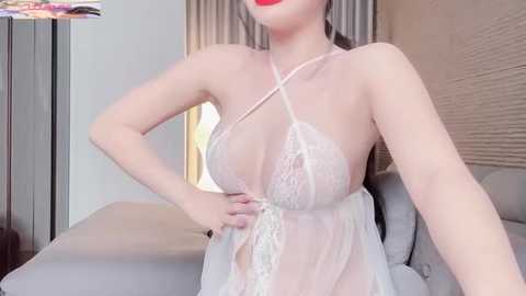 Media: Video of a fair-skinned woman in sheer white lace lingerie, featuring a halter top with a crisscross design, revealing her breasts. She stands in a modern bedroom with beige walls, a bed, and a partially visible picture frame.