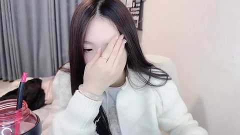 Media: Video of an East Asian woman with long black hair, wearing a white sweater, covering her face with her left hand, sitting in a chair with a pink pen and black notebook on the table in a room with grey curtains.