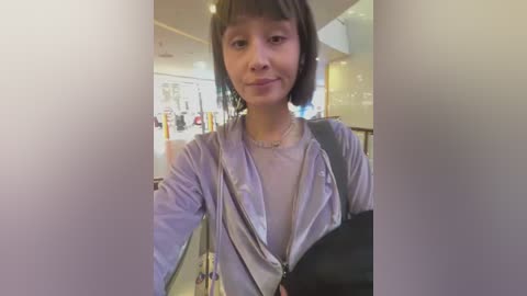 Media: Video of a young Asian woman with short dark hair, wearing a light purple zip-up jacket over a white shirt, smiling slightly. She carries a black bag, standing in a modern, well-lit indoor space with abstract art and white walls.