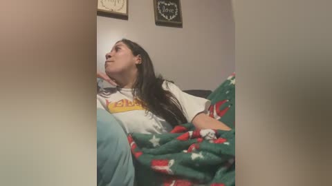 Media: Video of a young woman with long dark hair, wearing a white t-shirt with a yellow graphic, lying on a bed with a green blanket featuring Christmas-themed patterns. The background shows a neutral-toned room with framed wall art.