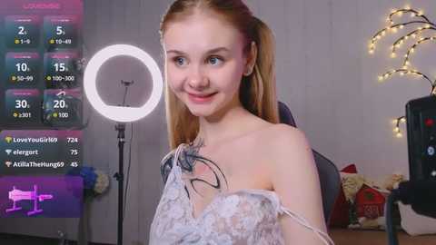 Media: Video of a young woman with fair skin, pigtails, and a delicate lace dress, smiling in a cozy, softly lit room with a ring light and a festive backdrop.