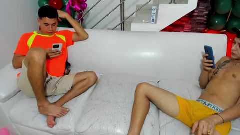 Media: Video of two shirtless young men with tanned skin, one in a bright orange shirt, one in yellow shorts, sitting on a white couch, looking at smartphones.