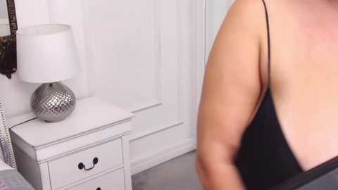 Media: Video of a woman with fair skin and large breasts in a black lace bra, standing next to a white nightstand with a silver lamp and white curtains in a modern, minimalist bedroom.