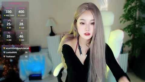 Media: Video of an East Asian woman with long, straight, platinum-blonde hair, wearing a black off-shoulder top, sitting in a white chair in a modern, dimly lit room.