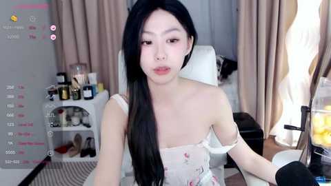 Media: A video of an Asian woman with long black hair, fair skin, and a slender physique, wearing a floral off-shoulder top, seated in a modern, cluttered room with a white chair, beige curtains, and a TV showing a live stream.