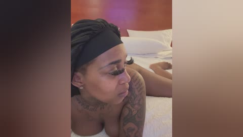 Media: Video of a dark-skinned woman with a black headscarf, tattoos on her arms, lying on a white bed in a dimly lit room.