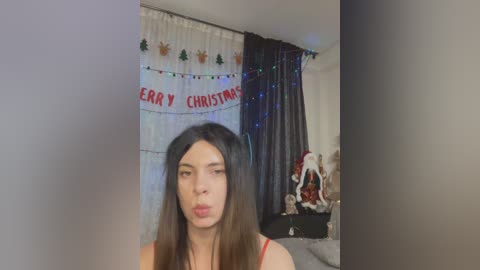 Media: Video of a young woman with long dark hair, wearing a red tank top, making a kissy face, against a festive Christmas-themed backdrop with \"Merry Christmas\" banner and Santa doll.