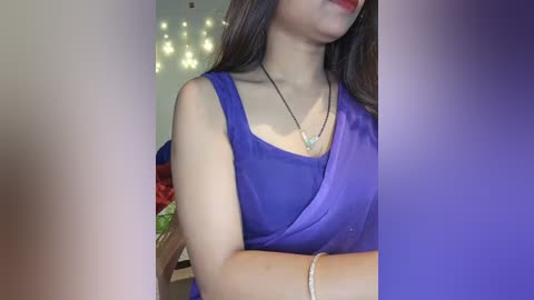 Media: Video of a woman in a vibrant purple saree, with a black necklace, silver bracelet, and long brown hair, set against a blurred, softly lit background.