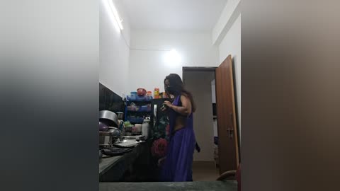 Media: Video of a narrow, dimly lit kitchen with a woman in a purple sari, holding a phone, reflected in a mirror. Shelves above the sink hold colorful pots and pans.