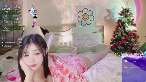 Media: A photo of a young East Asian woman with straight black hair, wearing a pink floral dress, lying on a bed. A Christmas tree with red ornaments is visible in the background.