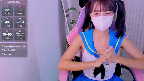 Media: Video of a young Asian woman in a pink gaming chair, wearing a white mask, blue sailor outfit, and glasses, displaying a health monitor on the left.