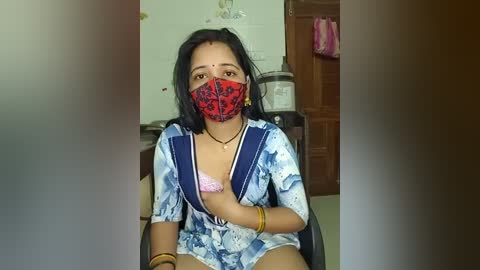 Media: Video of a woman in a blue tie-dye dress, red mask, and pink bra, sitting in a room with a wooden door, laundry, and a sink in the background.