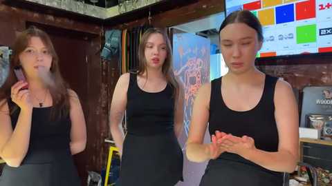 Media: Video of three young women in black dresses, holding hands, in a cluttered, dimly lit room with colorful posters on the wall and a television.