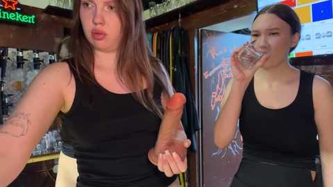 Media: Video of a light-skinned woman with long brown hair, wearing a black tank top, holding a clear plastic penis-shaped straw, standing in a bar with neon signs and shelves.