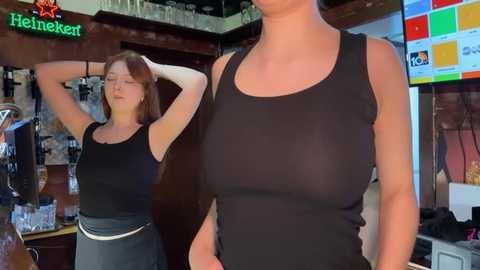 Media: A video of two women in a bar; one with large breasts in a see-through black tank top, the other with smaller breasts in a black top.