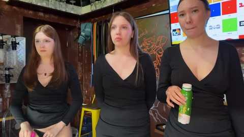 Media: Video of three young women in black V-neck sweaters, one holding a green drink, in a dimly lit, cozy bar with wooden walls and colorful TV screens.