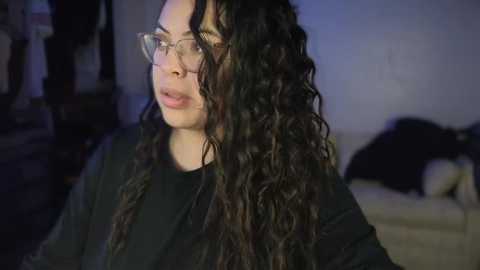 Media: Video of a young woman with long, wavy black hair and fair skin, wearing glasses, a black shirt, and looking slightly to the side. Background shows a dimly lit living room with a beige sofa and indistinct furniture.