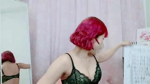 Media: Video of a young woman with short, vibrant red hair, wearing a black lace bra, adjusting a fan in a room with light pink curtains and a white mirror.