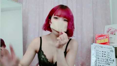 Media: Video of a young woman with bright red bob haircut, wearing a black lace bra, covering her mouth with a yellow plastic sheet. She stands in front of a mirror and a pink curtain, with a \"CENSORED\" sign on a whiteboard.