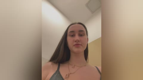 Media: Video of a young Caucasian woman with straight brown hair, wearing a gray tank top and gold necklace, standing in a narrow, dimly lit corridor with beige walls and a ceiling light.