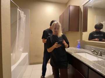 Media: Video of a blonde woman in a black shirt holding a phone, standing in a modern bathroom with a granite countertop, wooden cabinets, and a mirror.