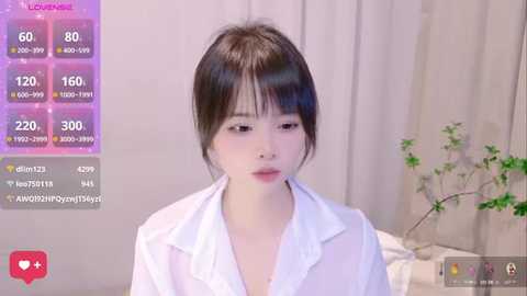 Media: A young East Asian woman with fair skin, black hair, and bangs, wearing a white blouse, stands in a softly lit room with beige curtains and a plant. The image features a live streaming overlay with heart icons and viewer counts.