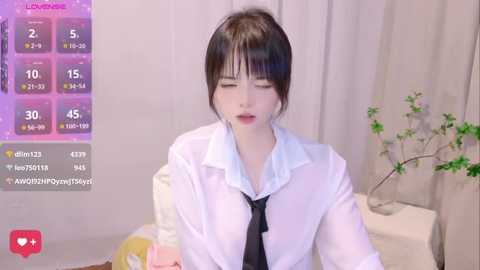 Media: Video of a young Asian woman with short black hair, wearing a white shirt and black tie, appearing seductive, in a modern bedroom with a plant.