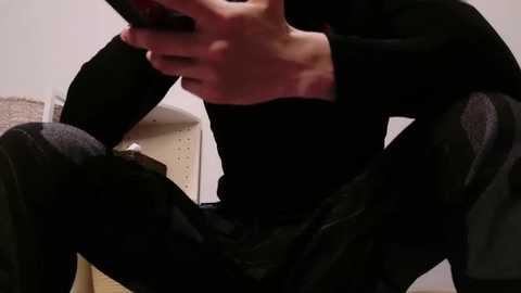 Media: Video of a person sitting on a chair, wearing a black long-sleeve shirt and dark pants, holding a smartphone, with a beige rug and a light-colored wall in the background.