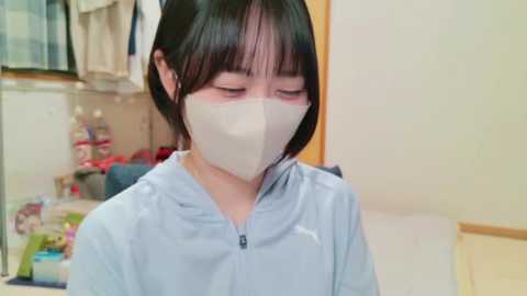 Media: Video of an Asian woman with short black hair and light skin, wearing a light blue scrub top and a beige face mask, looking down. Background shows a cluttered, beige-colored room with shelves, a sink, and hanging clothes.